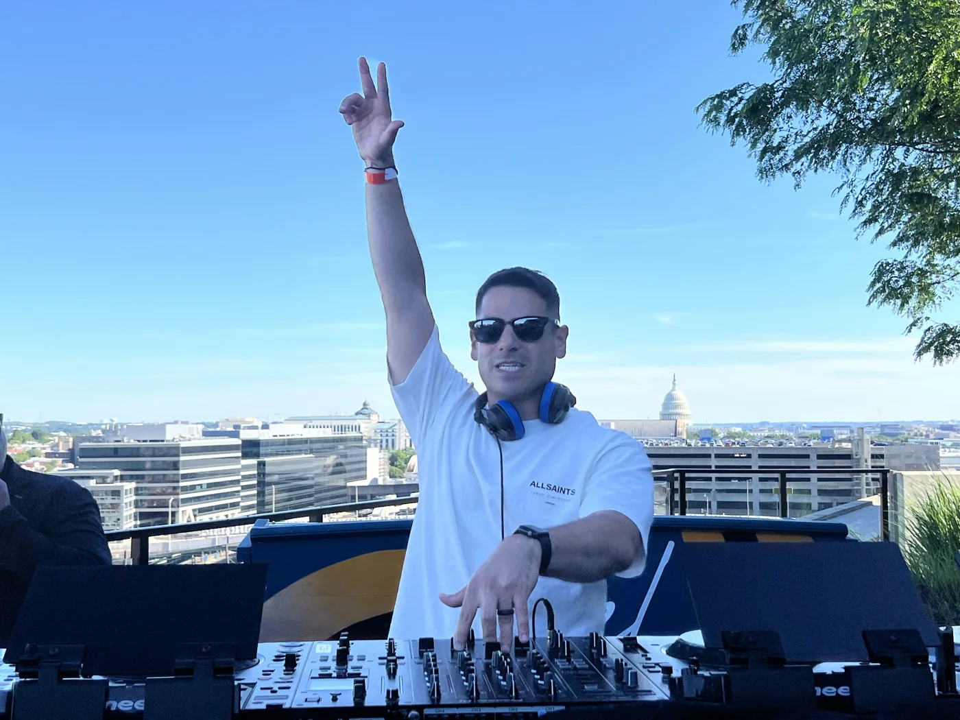 Andrew Weiss raising a hand behind the decks at a sunny rooftop gig
